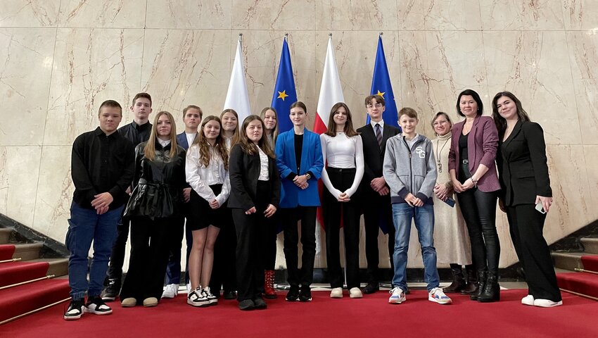 Powiększ: Grupa młodzieży z nauczycielkami na tle flag w gmachu budynku